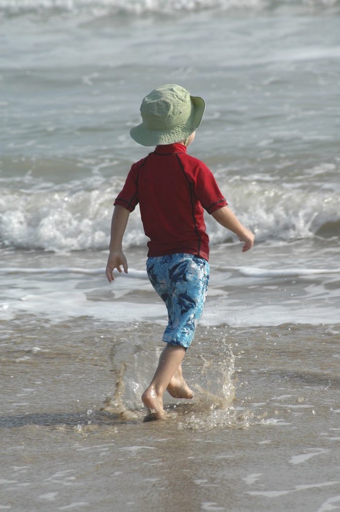 A young boy running in the ocean with a hat on.