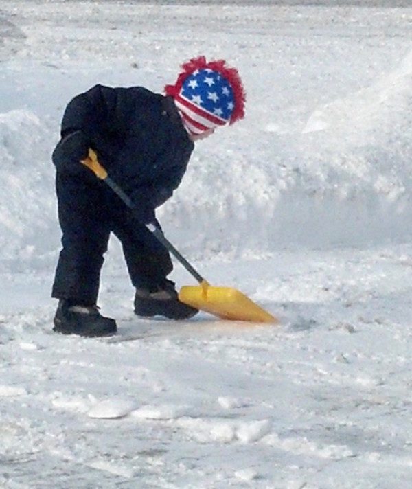 A boy wearing an american flag hat shoveling snow.